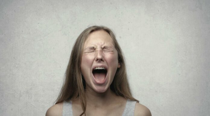 Как научиться проговаривать эмоции без страха и вины Young woman screaming with emotion, showing braces, against a gray backdrop.