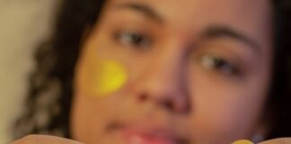 Soft focus of African American young female demonstrating eye patch in hands and looking at camera during skincare routine on beige background